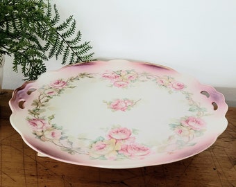 Puede incluir: Un plato de pastel de porcelana blanca vintage con un borde degradado rosa y morado. El plato está decorado con un patrón floral de rosas rosas y hojas verdes.