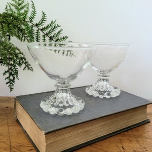 May include: Two clear glass stemmed dessert cups with a beaded base, sitting on a closed book.