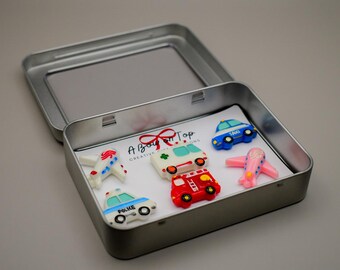 May include: A metal tin containing toy vehicles, including a blue taxi, a red fire truck, and a pink airplane. The tin also contains a white ambulance, a white police car, and a white airplane. The text "A Boy on Top" is visible.