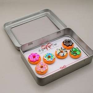 Puede incluir: Una caja plateada abierta que contiene seis imanes en forma de mini rosquillas coloridas. Cada imán tiene un diseño diferente de glaseado y chispas. El interior de la caja presenta una tarjeta blanca con el texto "A Bow on Top".