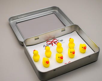 Puede incluir: Una lata de plata con fondo blanco y una cinta roja. La lata contiene nueve figuras de patos de goma amarillos con picos rojos.