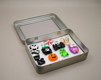 Puede incluir: Una lata de metal plateado que contiene una colección de pequeños dijes de resina con temática de Halloween. Los dijes incluyen una calavera, un gato negro, un caldero, una calabaza, un murciélago, un fantasma y un búho. El texto A Bow on Top es visible.
