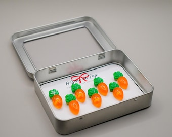 Puede incluir: Una lata plateada abierta que contiene varios caramelos con forma de zanahoria de color naranja con tapas verdes. Los caramelos están sobre una tarjeta blanca con el texto "A Bow on Top". La lata está sobre una superficie gris claro.