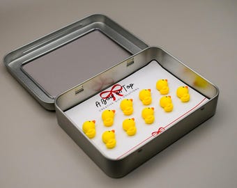 Puede incluir: Una lata de plata con una tarjeta blanca en su interior. La tarjeta tiene el texto "A Bow on Top Creative Gift Solutions" y presenta 12 figuras de patos de goma amarillos.