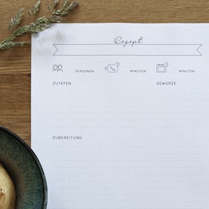 Könnte beinhalten: Ein weißes Rezeptblatt mit dem Wort "Rezept" oben, mit Abschnitten für Zutaten, Zubereitung und Garzeiten. Eine Schüssel mit Zimtschnecken und ein Zweig Grünzeug liegen auf einer Holzoberfläche.