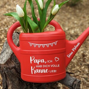 May include: A red watering can with white text and a banner design, holding white tulips. The text on the can reads "Papa, ich hab dich volle Kanne lieb!" and "Deine Melanie". The can is sitting on a tree stump.