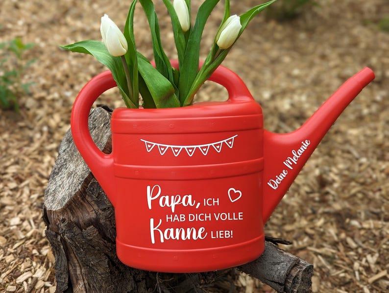 May include: A red watering can with white text and a banner design, holding white tulips. The text on the can reads "Papa, ich hab dich volle Kanne lieb!" and "Deine Melanie". The can is sitting on a tree stump.