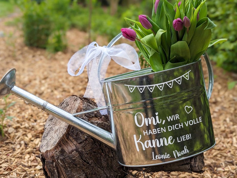 Könnte beinhalten: Eine verzinkte Metall-Gießkanne gefüllt mit lila Tulpen, die auf einem Baumstumpf ruht. Die Kanne hat ein weißes Band am Griff und weißen Text, der "Omi, wir haben dich volle Kanne lieb!" mit einem Herzen und dem Namen "Leonie Noah" lautet.