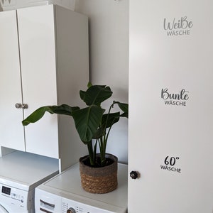 Può includere: Armadi bianchi per lavanderia con pomelli decorativi. Le ante degli armadi hanno etichette di testo in tedesco: "Weiße Wäsche", "Bunte Wäsche" e "60° Wäsche". Una pianta in vaso in un cestino intrecciato si trova tra gli armadi e una lavatrice.
