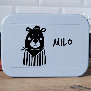 Op de afbeelding: Een lichtblauwe rechthoekige lunchbox met een zwarte grafische print van een beer met een hoed en een halsdoek, met de naam Milo in zwarte letters rechts van de beer. De container is voorzien van zwarte sluitingen aan de zijkanten.