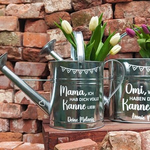 May include: Two galvanized metal watering cans with white text and floral arrangements. The text on the first can reads "Mama, ICH HAB DICH VOLLE Kanne LIEB! Deine Melanie." The second can reads "Omi, WIR HABEN DICH VOLLE Kanne LIEB! Leonie Noah."