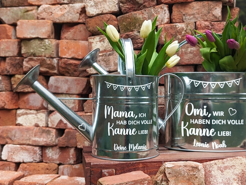 May include: Two galvanized metal watering cans with white text and floral arrangements. The text on the first can reads "Mama, ICH HAB DICH VOLLE Kanne LIEB! Deine Melanie." The second can reads "Omi, WIR HABEN DICH VOLLE Kanne LIEB! Leonie Noah."