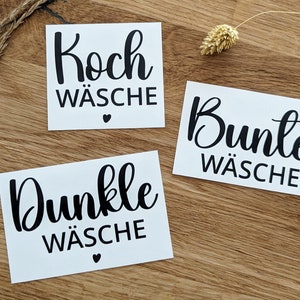Peut inclure: Trois étiquettes rectangulaires blanches avec du texte noir sur une surface en bois. Les étiquettes indiquent "Koch Wäsche", "Bunte Wäsche" et "Dunkle Wäsche". Chaque étiquette a un petit symbole de cœur noir.
