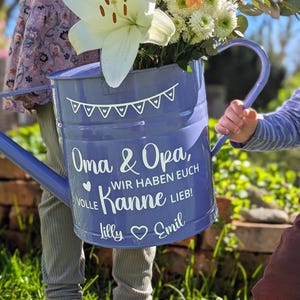 May include: A blue watering can filled with white and orange flowers. The can has a white banner design and text that reads "Oma & Opa, WIR HABEN EUCH VOLLE Kanne LIEB!" with names "Lilly" and "Emil".