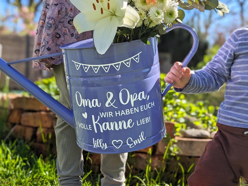 May include: A blue watering can filled with white and orange flowers. The can has a white banner design and text that reads "Oma & Opa, WIR HABEN EUCH VOLLE Kanne LIEB!" with names "Lilly" and "Emil".