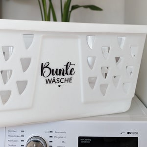 Op de afbeelding: Een witte plastic wasmand met driehoekige uitsparingen. De woorden "Bunte Wäsche" zijn in zwarte letters op de zijkant gedrukt. De mand staat op een witte wasmachine.