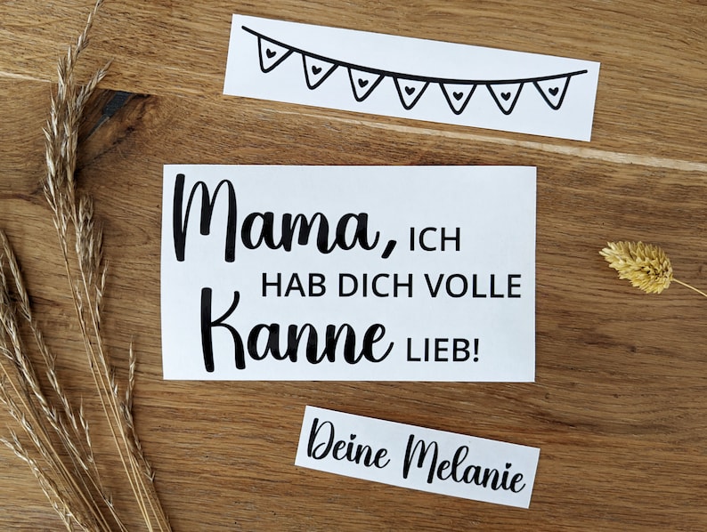 May include: White sticker with the text "Mama, ICH HAB DICH VOLLE Kanne LIEB!" in black script. A smaller sticker reads "Deine Melanie". A banner with heart-shaped flags is also included. The stickers are on a wooden surface.