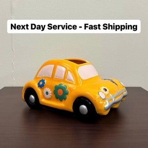 May include: A yellow ceramic car-shaped planter with black wheels and white accents. The car is decorated with blue, white, and green flower designs. The planter sits on a dark brown surface.