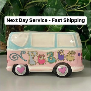 May include: A ceramic planter shaped like a vintage van, painted pink, blue, and white. The word "Peace" is written on the side in colorful letters. The wheels have heart-shaped centers. Decorative item.
