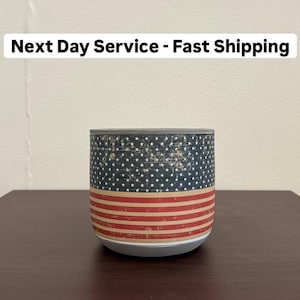 May include: Cylindrical object with a design resembling the American flag. The top portion features a dark blue background with white stars, while the bottom has red and white stripes. The text "Next Day Service - Fast Shipping" is above the object.