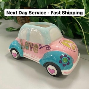 May include: A ceramic car-shaped planter in pastel pink, blue, and white. The word "Love" is written on the side, and a peace sign is on the roof. The wheels have heart details, and a flower decorates the fender.