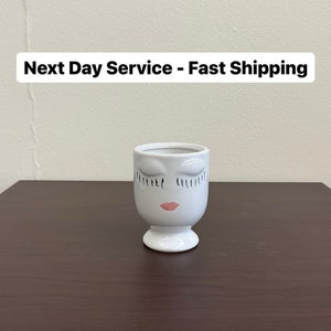 May include: White ceramic planter shaped like a face with closed eyes and pink lips. The planter is on a dark brown wooden surface.