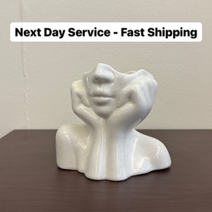 May include: A white ceramic face-shaped vase with a glossy finish. The vase depicts a face with hands supporting the chin. The top of the vase is open for flowers or other decorative items. The vase is on a dark brown surface.