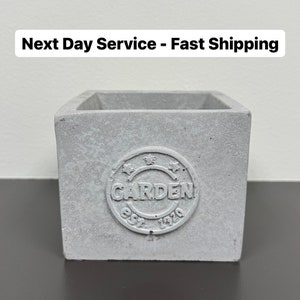 May include: A square, light gray concrete planter with the word "GARDEN" in a circular emblem. The emblem also includes the text "EST. 1979". The planter is empty and sits on a dark surface.