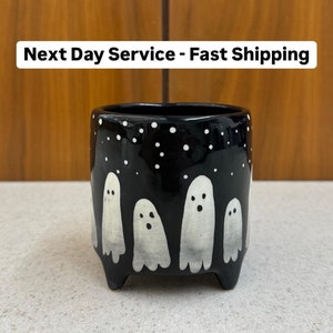 May include: Black ceramic planter with a whimsical ghost design. The planter features white ghost figures against a black background with white dot accents. The planter has a square shape and is elevated by small feet.