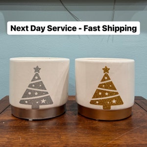 May include: Two white ceramic candle holders with a gold and silver glitter Christmas tree design. The candle holders are on a wooden surface. "Next Day Service - Fast Shipping" is written on a sign above the candle holders.