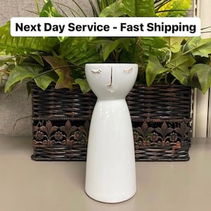 May include: White ceramic vase with a stylized face design, featuring closed eyes and a gold nose and mouth. The vase is set against a backdrop of greenery and a decorative basket. The text "Next Day Service - Fast Shipping" is visible.