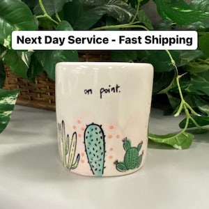 May include: A white ceramic mug with a hand-drawn cactus design in green, blue, and pink. The mug has the words "on point" written on it. The background includes green plants.