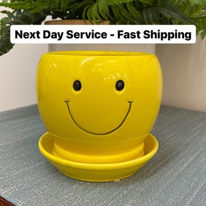May include: A yellow ceramic smiley face planter with a matching saucer, filled with green leafy plants. The planter has black eyes and a curved smile. The text "Next Day Service - Fast Shipping" is at the top.