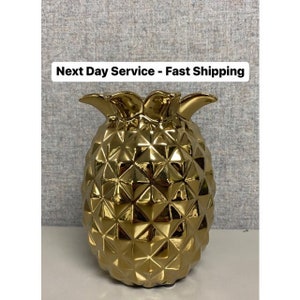 May include: A gold-colored pineapple-shaped decorative object. The pineapple has a textured surface with a geometric pattern. The top of the pineapple has a crown of leaves. The object is on a white surface.