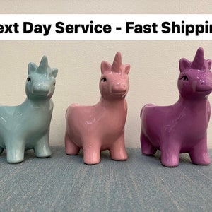 May include: Three ceramic unicorn planters in pastel colors: light blue, pink, and purple. Each unicorn has a horn and a small opening on its back for planting small succulents or flowers. The planters are displayed on a blue surface.