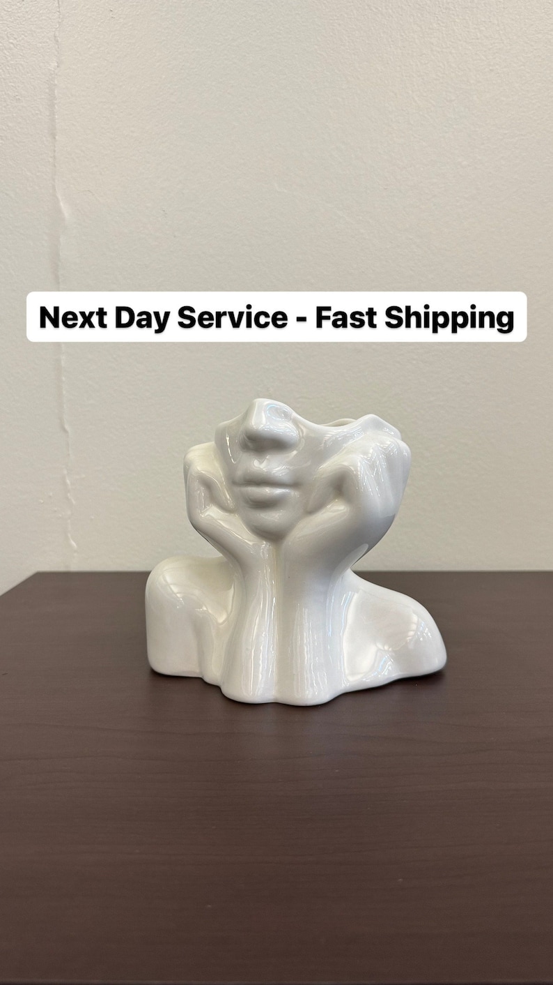 May include: A white ceramic face-shaped vase with a glossy finish. The vase depicts a face with hands supporting the chin. The top of the vase is open for flowers or other decorative items. The vase is on a dark brown surface.