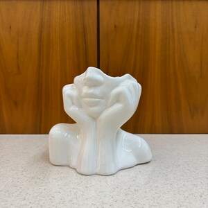 May include: A white ceramic decorative vase shaped like a face with hands supporting the head. The vase has a smooth, glossy finish and is placed on a light-colored surface. The background features a wooden cabinet.