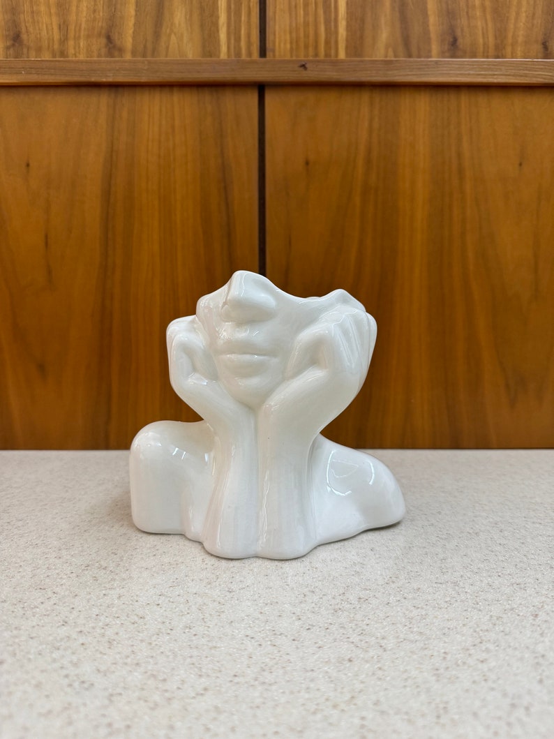May include: A white ceramic decorative vase shaped like a face with hands supporting the head. The vase has a smooth, glossy finish and is placed on a light-colored surface. The background features a wooden cabinet.
