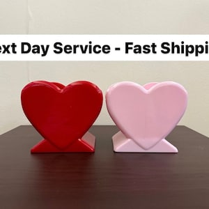 May include: Two ceramic heart-shaped planters, one red and one pink. The planters are sitting on a dark brown surface.