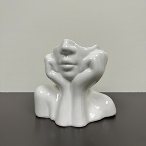 May include: A white ceramic face sculpture with hands supporting the chin. The sculpture has a smooth, glossy finish and is set against a neutral background. The face features a defined nose and lips, with the top of the head open.
