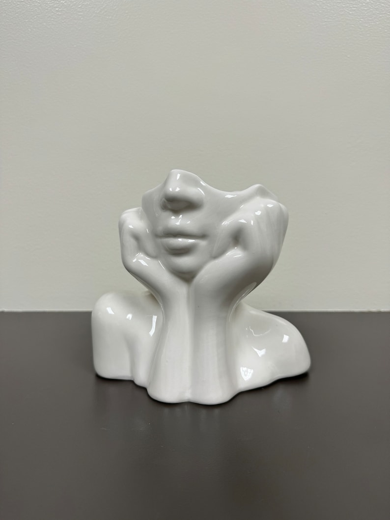 May include: A white ceramic face sculpture with hands supporting the chin. The sculpture has a smooth, glossy finish and is set against a neutral background. The face features a defined nose and lips, with the top of the head open.