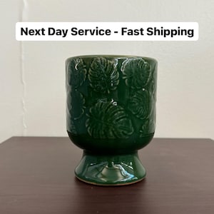 May include: A green ceramic planter with a raised leaf design. The planter has a wide base and a narrow top.