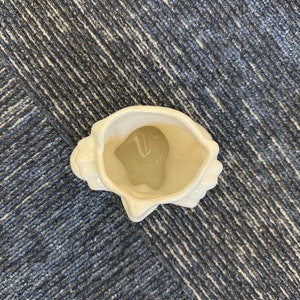 May include: A white ceramic seashell-shaped decorative vase. The vase has a glossy finish and a detailed, textured exterior, with a smooth interior. The opening is wide, and the overall design is reminiscent of a natural seashell. The vase is empty.