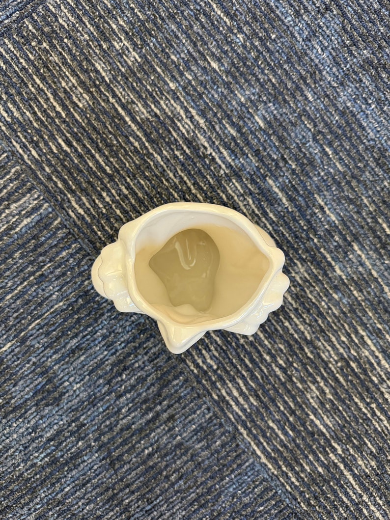May include: A white ceramic seashell-shaped decorative vase. The vase has a glossy finish and a detailed, textured exterior, with a smooth interior. The opening is wide, and the overall design is reminiscent of a natural seashell. The vase is empty.