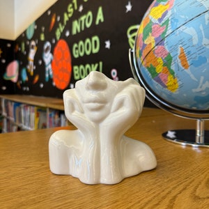 May include: A white ceramic decorative piece shaped like a face with hands supporting the head. The piece is on a wooden surface, with a globe and a wall with the text "BLAST OFF INTO A GOOD BOOK!" in the background.