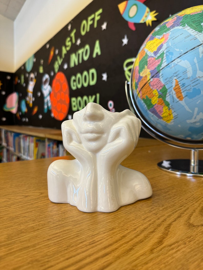 May include: A white ceramic decorative piece shaped like a face with hands supporting the head. The piece is on a wooden surface, with a globe and a wall with the text "BLAST OFF INTO A GOOD BOOK!" in the background.
