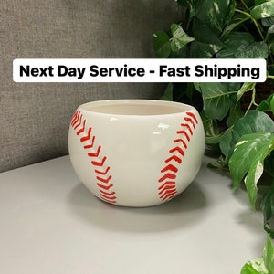 May include: A white ceramic baseball-themed planter with red stitching detail. The planter is round and has an open top. The text "Next Day Service - Fast Shipping" is displayed above the planter.