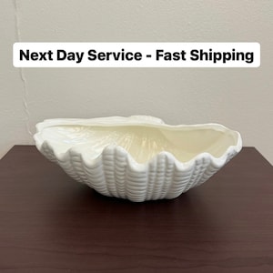 May include: White ceramic seashell-shaped bowl. The bowl is on a dark brown wooden surface.