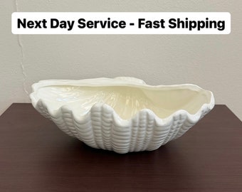 May include: White ceramic seashell-shaped bowl. The bowl is on a dark brown wooden surface.