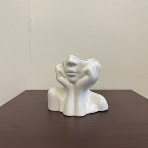 May include: A white ceramic vase shaped like a human face, with hands supporting the head. The vase has a smooth, glossy finish and sits on a dark brown surface. The face has a serene expression.